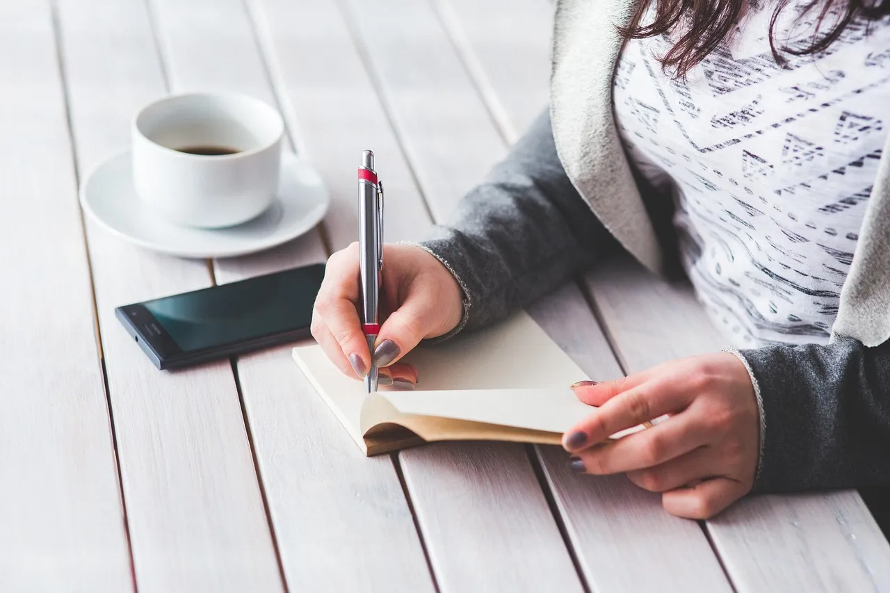 Woman writing in a notebook with a cup of coffee and smartphone on a wooden table.