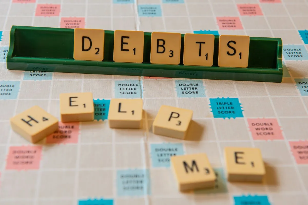Scrabble tiles spell "DEBTS" and "HELP ME" on a game board.