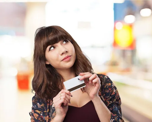 Young woman holding a blank credit card, looking thoughtfully in a blurred shopping environment.