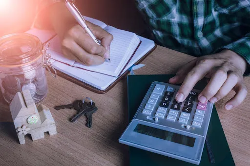 Person calculating expenses with a calculator, notebook, and coins on a wooden table.