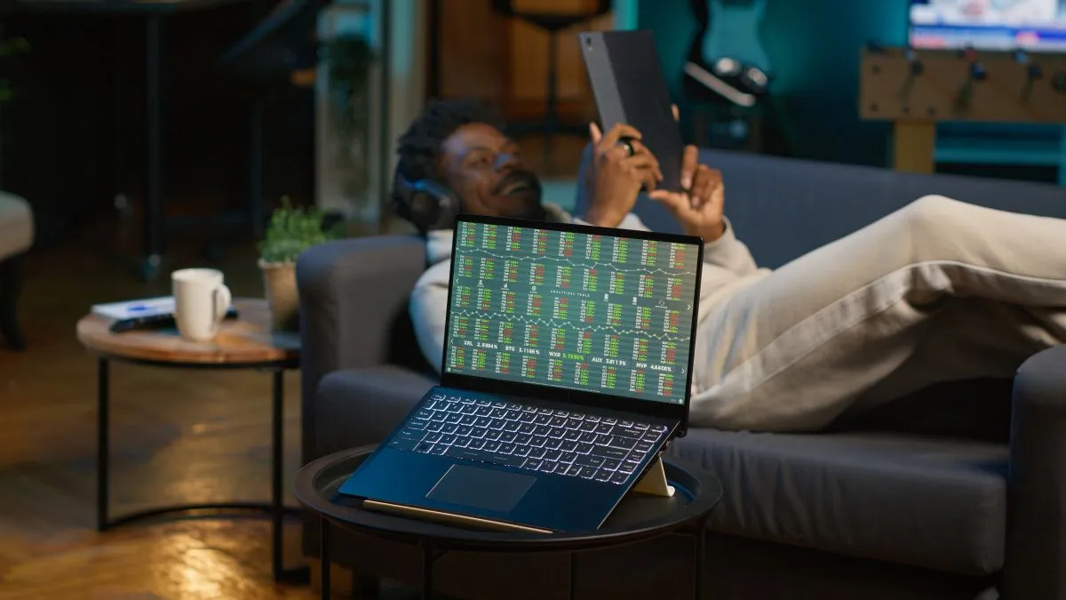 A person relaxing on a couch, using a tablet while a laptop displays stock market data.