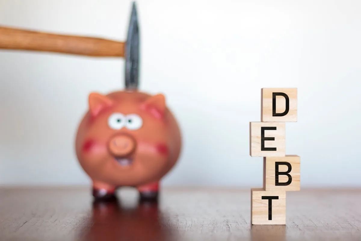 Piggy bank with a hammer behind it, wooden blocks spelling "DEBT" in focus.