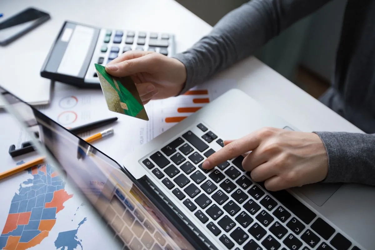 Persona usando una laptop para hacer un pago en línea con una tarjeta de crédito, rodeada de documentos financieros y una calculadora.