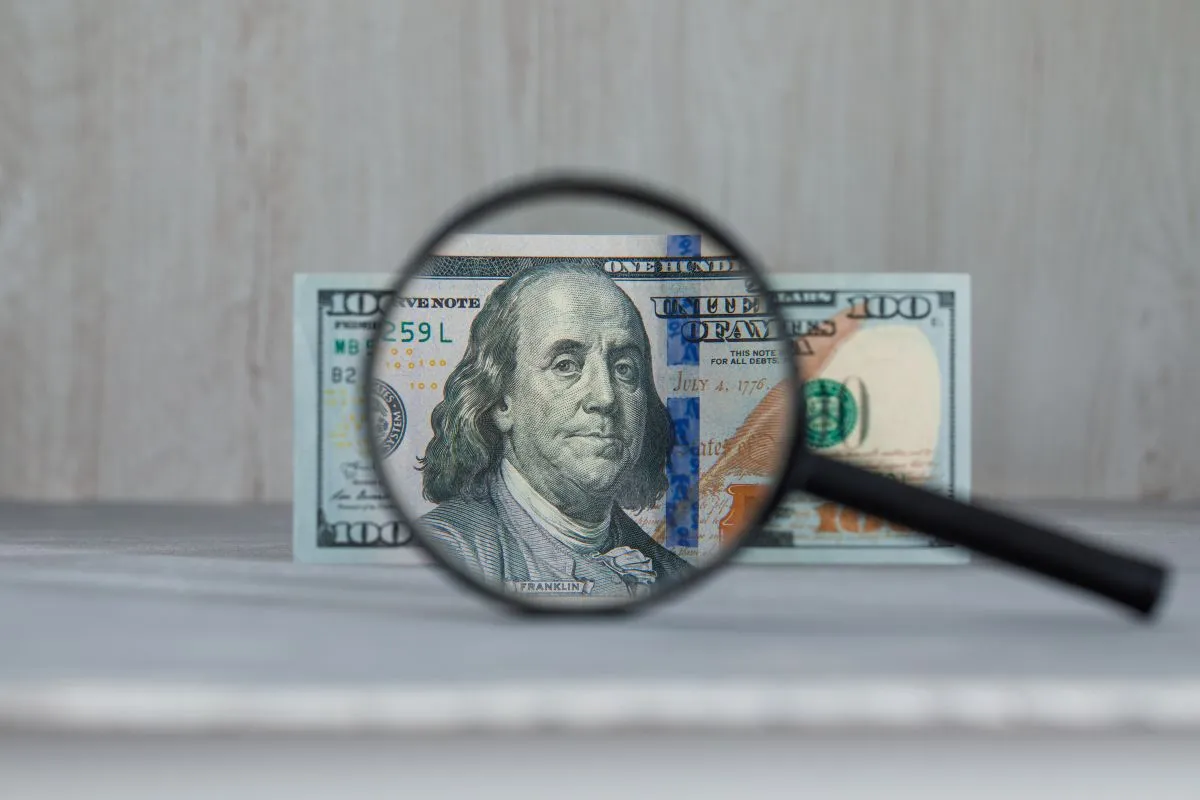 Magnifying glass focusing on a U.S. hundred-dollar bill featuring Benjamin Franklin.