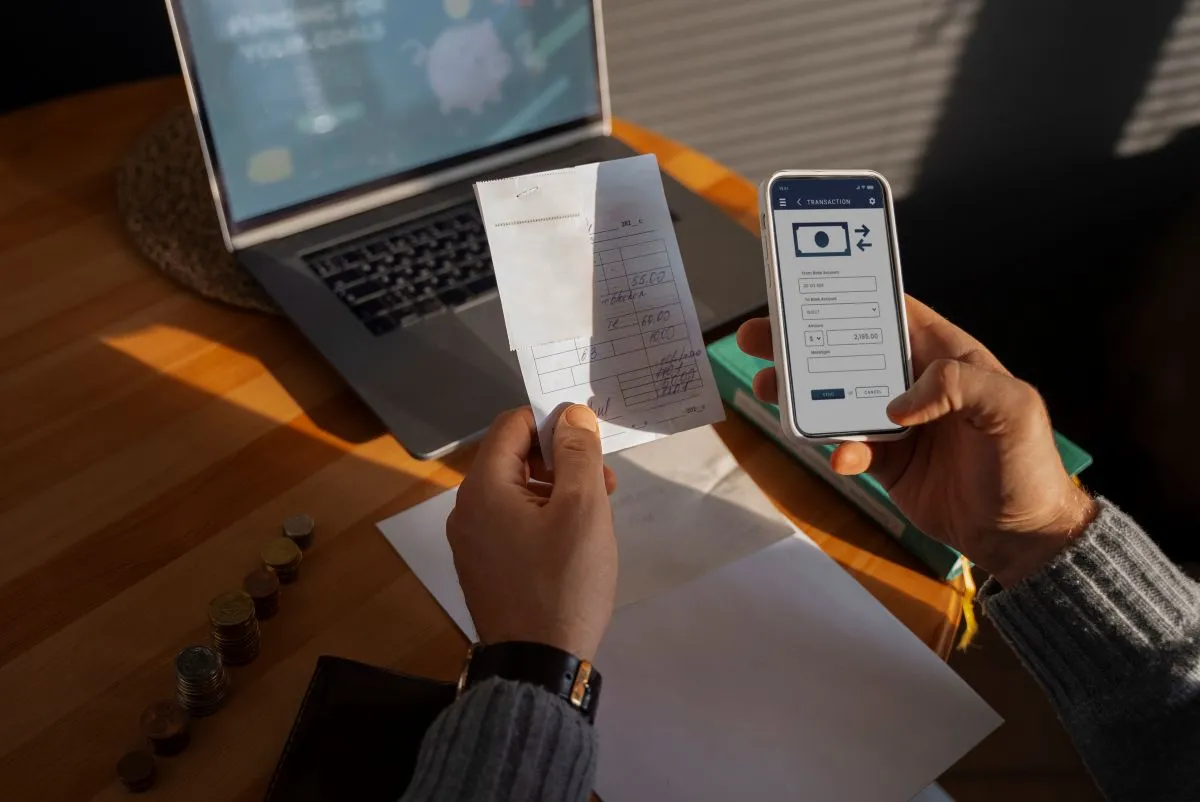 Person comparing a receipt with a mobile banking app on a smartphone, surrounded by coins and a laptop.