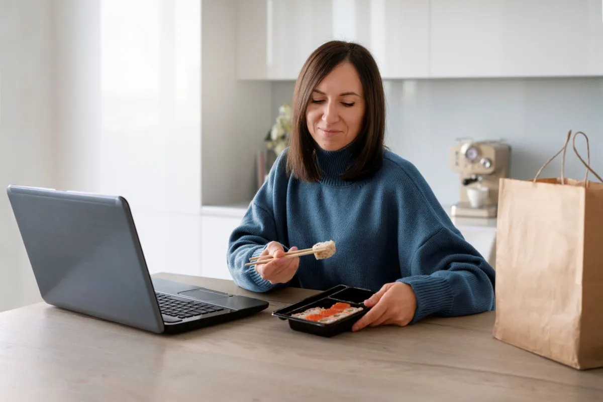 Woman enjoying sushi while working on a laptop at home, with a takeout bag nearby.