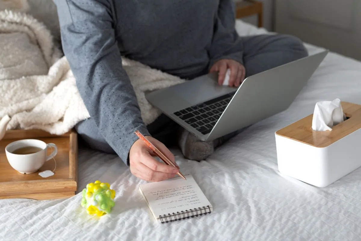 Cozy home workspace with a person writing notes, using a laptop, coffee, and tissues nearby.