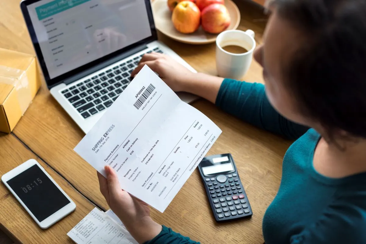 Woman reviewing an invoice while using a laptop, calculator, and smartphone, with apples and coffee on the table.
