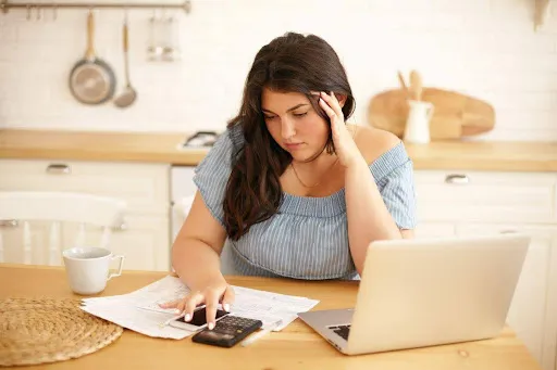 Young woman calculating expenses at a kitchen table with a laptop and papers.