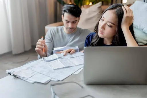 Couple reviewing bills and financial documents at home, looking stressed and concerned.