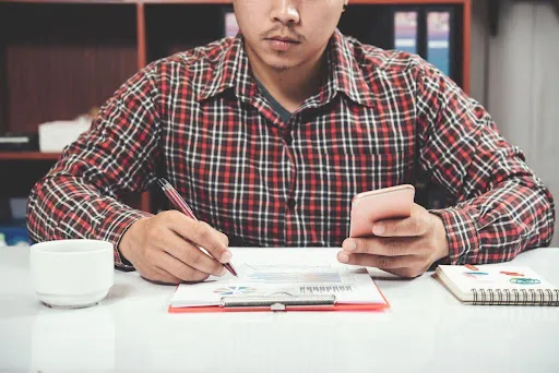 Man in a plaid shirt analyzing charts while taking notes and using a smartphone at a desk.
