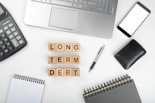 Calculator, laptop, smartphone, and notebooks surrounding wooden blocks spelling "LONG TERM DEBT."