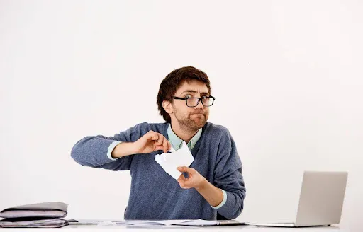 Frustrated man shredding paperwork at a cluttered desk with a laptop.