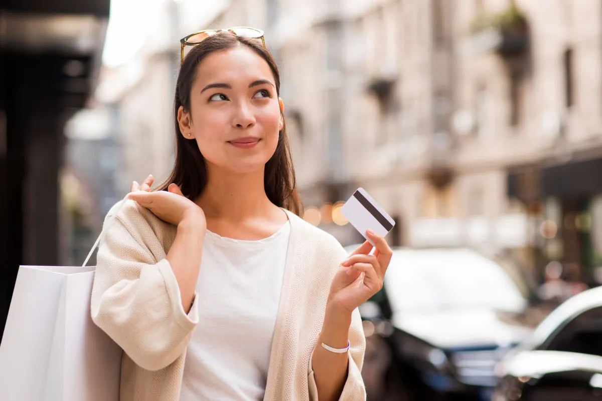 Young woman holding a credit card and shopping bag, smiling on a city street.