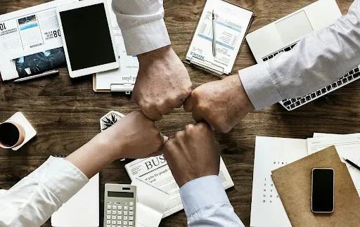Team members fist bumping over a wooden desk filled with business documents, a laptop, and office supplies.