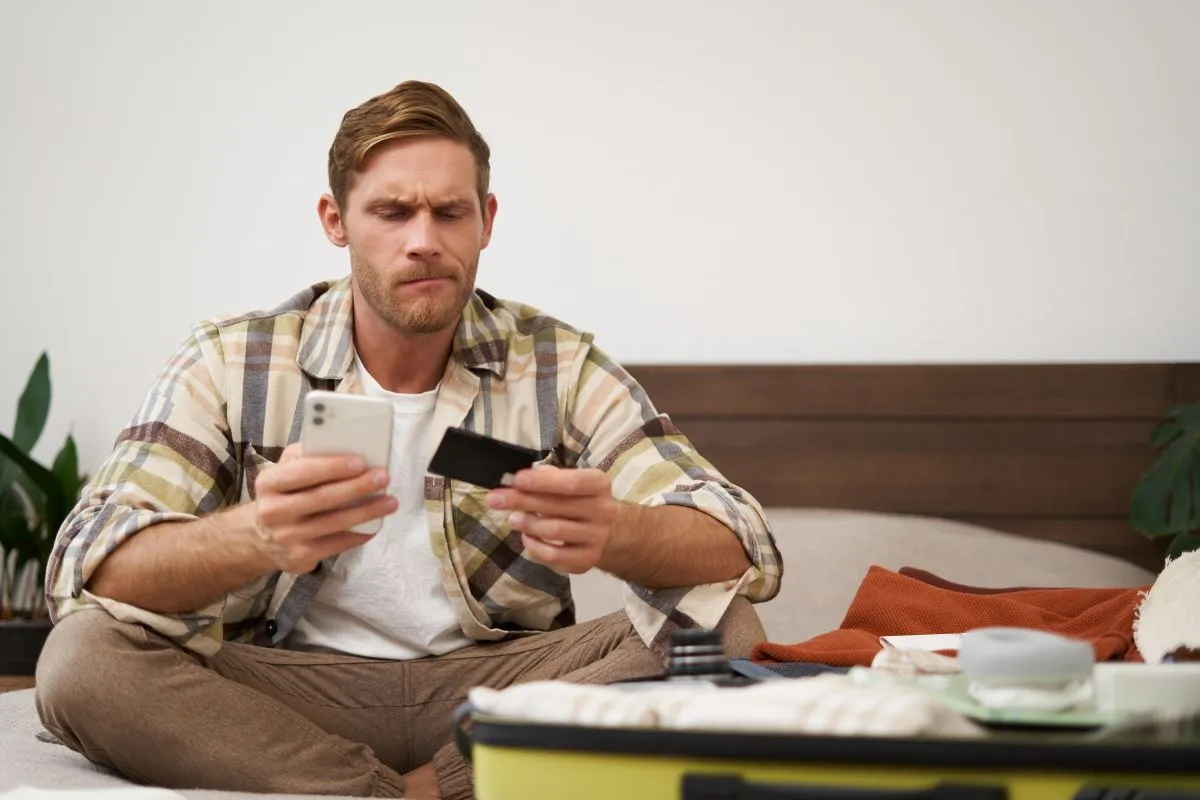 Man sitting on a bed, looking concerned while holding a smartphone and credit card, surrounded by luggage and travel items.
