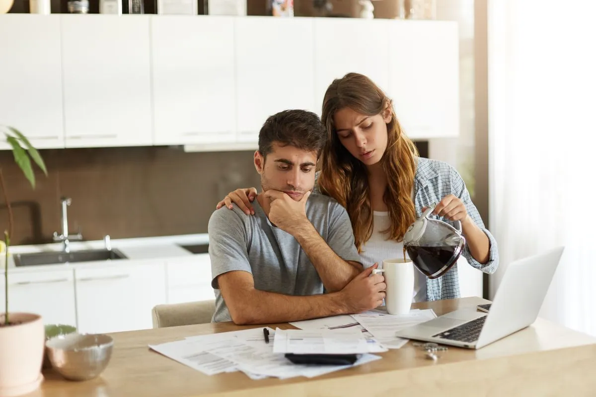 Couple discussing finances at home, with one pouring coffee while the other looks concerned.